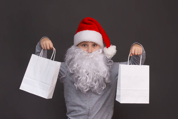 little Santa boy is holding two white paper bags in his hands. on a gray background.  - 写真・画像