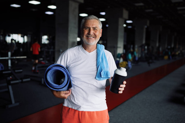 Γέρος με μπουκάλι νερό, πετσέτα και χαλάκι στο γυμναστήριο - Φωτογραφία, εικόνα