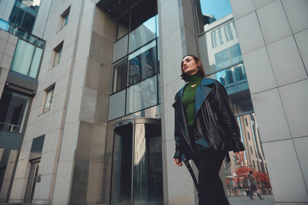 Femme brune à la mode avec une coupe de cheveux courte élégante marchant le long de la rue avec des bâtiments modernes de grande hauteur d'entreprise sur le fond. Portrait de style de vie - Photo, image