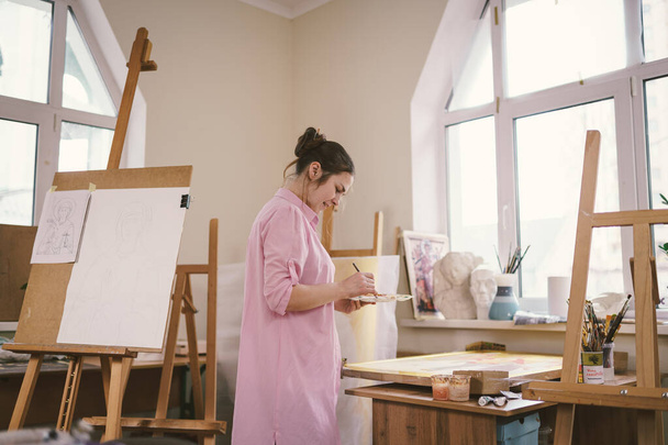 Caucasian woman artist working on a painting in bright daylight studio. Happy artist draws an art project with paints and a brush in the workshop. Hobby. Artist at work. Creative profession. - 写真・画像