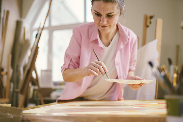 Cute woman paints on canvas in an art workshop. Artist creating picture. Art school or studio. Work with paints, brushes and easel. Hobby and leisure concept. Woman painter at workspace. - Photo, Image