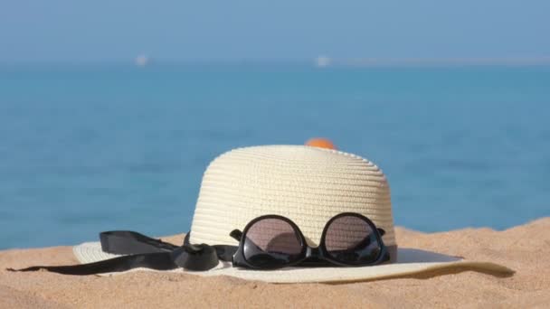 Szekrény sárga szalma kalap és fekete védő napszemüveg homokos strandon a trópusi tengerparton, meleg napsütéses napon. Nyári szabadság koncepciója - Felvétel, videó