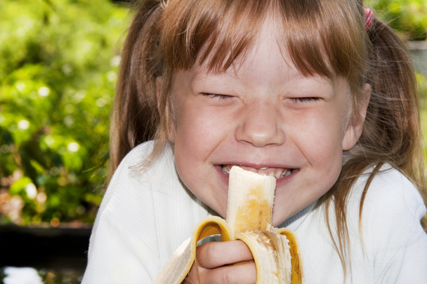 Little girl in the outdoors eats a banana and laughs - Zdjęcie, obraz