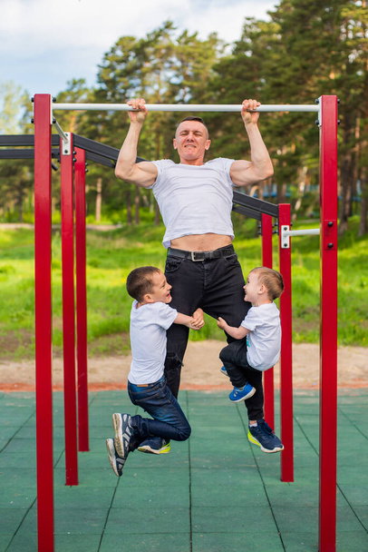 Caucasian man and two boys doing exercises outdoors. The father pulls himself up on the horizontal bar with his sons on the playground. - Фото, зображення