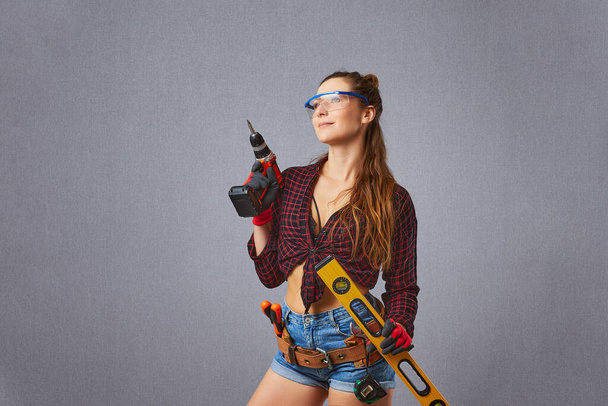 Young longhaired woman in casual clothes protective gloves hold electric drill or screwdriver in studio. Instruments accessories for renovation apartment room. - Φωτογραφία, εικόνα