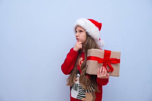 Una chica con el pelo largo en un sombrero de Santa Claus rojo sostiene su barbilla pensativamente, sosteniendo una caja con un regalo para Navidad y Año Nuevo en su mano. Lugar para la publicidad - Foto, Imagen