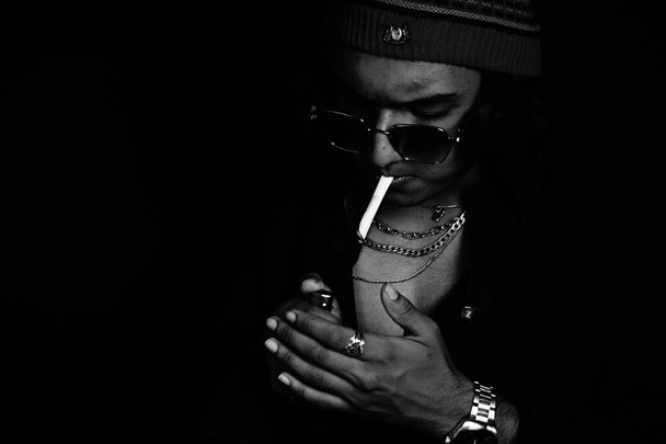 A grayscale shot of a stylish young man in sunglasses and jewelry posing in a dark room - Foto, imagen