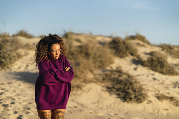 A sexy tattooed female in a long purple sweater posing on a beach - Zdjęcie, obraz