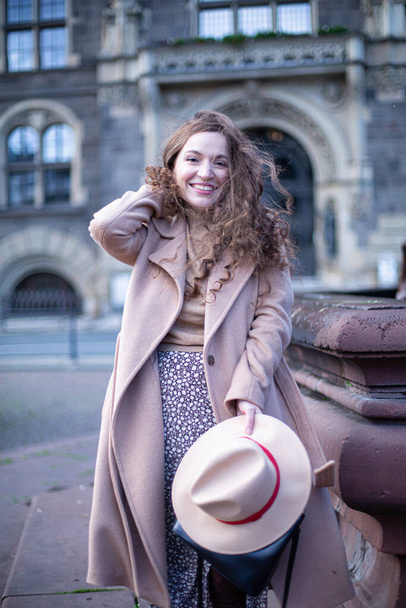 Smiling pretty woman with curly hair in autumn coat with old town buildings behind - Photo, image