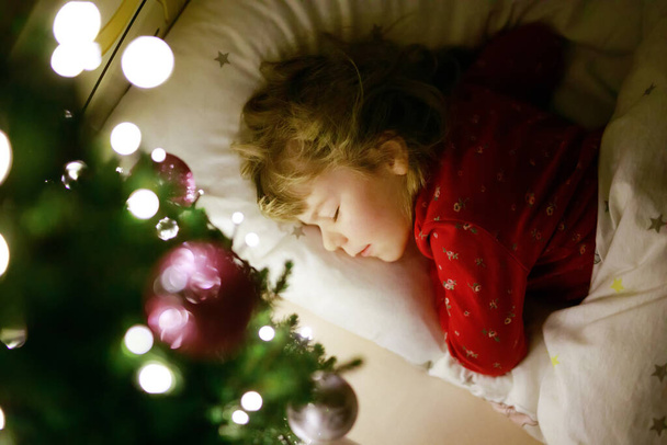 Kleine süße Mädchen schlafen unter dem Weihnachtsbaum und träumen vom Weihnachtsmann zu Hause, drinnen. Traditionelles christliches Fest. Glückliches Kind, das auf Geschenke zu Weihnachten wartet. Kuscheliges weiches Licht - Foto, Bild