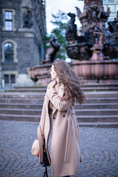 Looking away pretty woman with curly hair in autumn coat with old town buildings behind - Фото, изображение