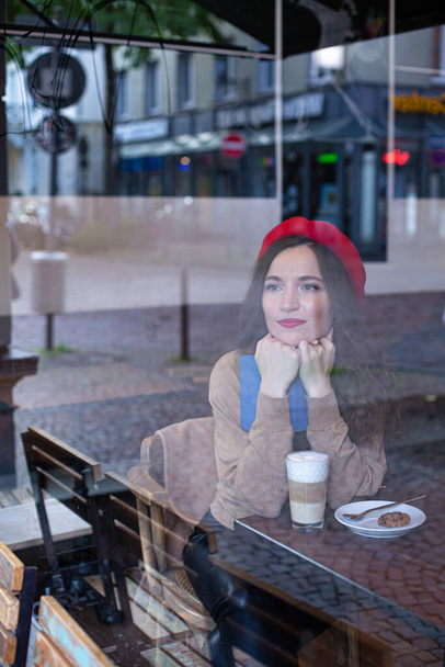 Pretty woman with curly hair in red french beret outside the cafe window reading the book with a cup of latte - Фото, изображение