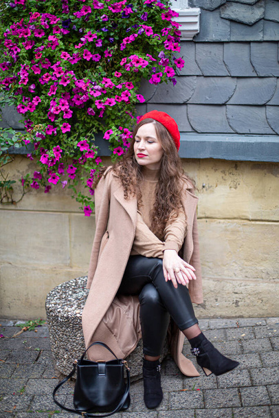 Pretty woman with curly hair in red french beret with building behind and petunia flowers - Foto, imagen