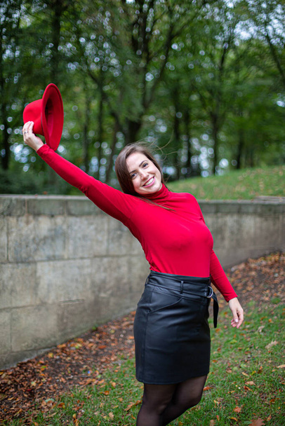 Smiling white woman with long dark hair in bright red near stone wall in autumn park holding her hat in right hand - Valokuva, kuva