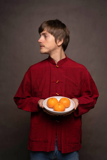Young handsome tall slim white man with brown hair holding a bowl of oranges in red shirt on grey background - Valokuva, kuva