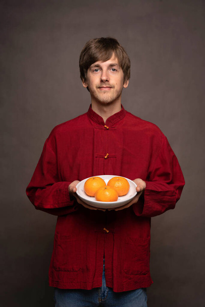 Young handsome tall slim white man with brown hair offering bowl of oranges in red shirt on grey background - Foto, immagini