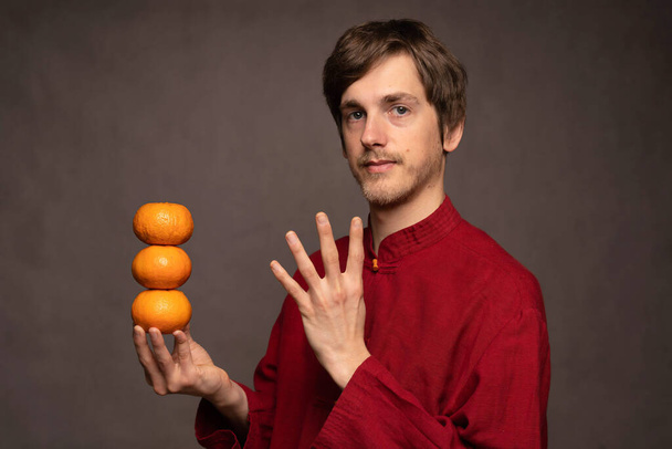Young handsome tall slim white man with brown hair calmly holding oranges in red shirt on grey background - Φωτογραφία, εικόνα