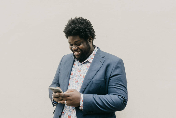 A shallow focus of a smiling black man looking at his phone on a blank wall background - Φωτογραφία, εικόνα