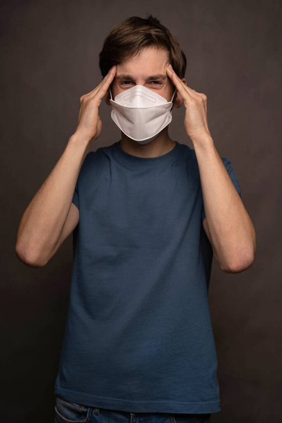 Young handsome tall slim white man with brown hair wearing face mask with his head hurting in blue shirt on grey background - Fotografie, Obrázek