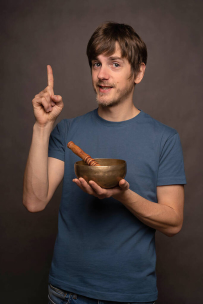 Young handsome tall slim white man with brown hair pointing up holding tibetan singing bowl in blue shirt on grey background - Zdjęcie, obraz