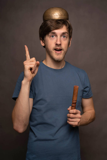 Young handsome tall slim white man with brown hair coming up with idea tibetan singing bowl on head in blue shirt on grey background - Фото, изображение