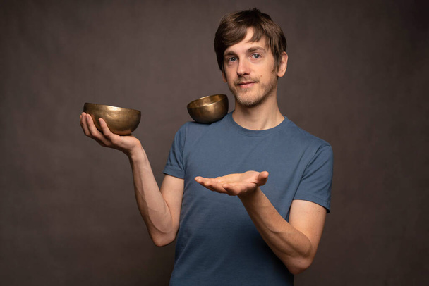 Young handsome tall slim white man with brown hair holding bowl while balancing another bowl on shoulder in blue shirt on grey background - Foto, Bild
