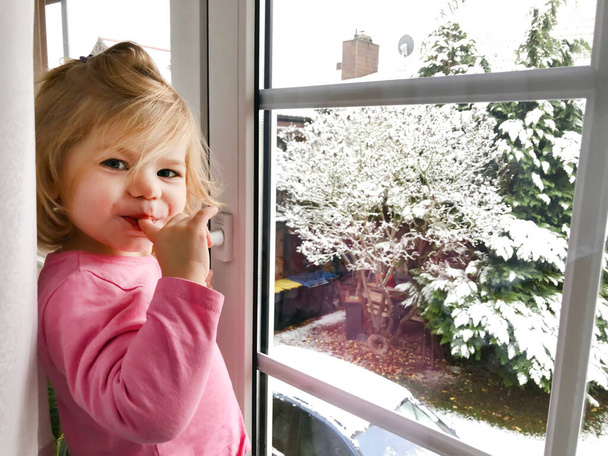 Счастливая очаровательная девочка, сидящая у окна и выглядывающая на улицу по снегу в день Рождества или утром. Улыбающийся здоровый ребенок восхищался снегопадами и большими снежинками. - Фото, изображение