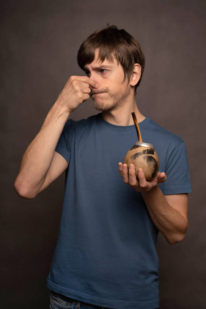 Young handsome tall slim white man with brown hair holding his nose holding mate cup in blue shirt on grey background - Fotografie, Obrázek