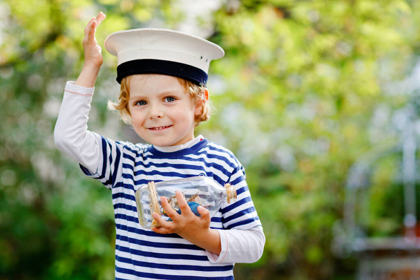 Happy little kid boy in sailor capitain hat and uniform playing with sailor boat ship. Smiling preschool child dreaming and having fun. Education, profession, dream concept - Photo, Image