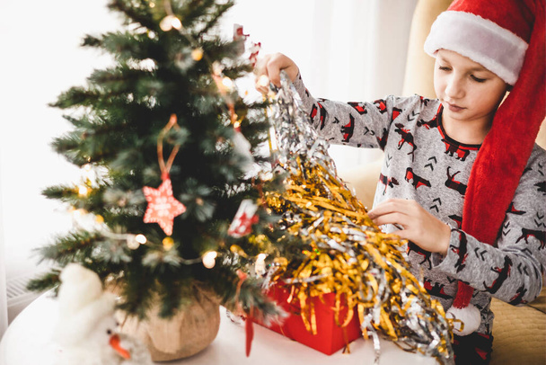 Święta i Nowy Rok. Szczęśliwy chłopiec w piżamie i czerwony kapelusz Mikołaja otwierający pudełko w domu wczesnym rankiem. Duch wakacji zimowych - Zdjęcie, obraz