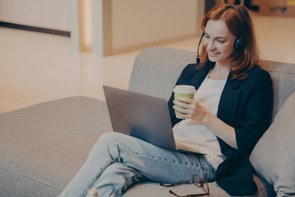 Usmívající se mladá žena call centra agent v bezdrátové sluchátka a notebook sedí na pohovce v kavárně - Fotografie, Obrázek