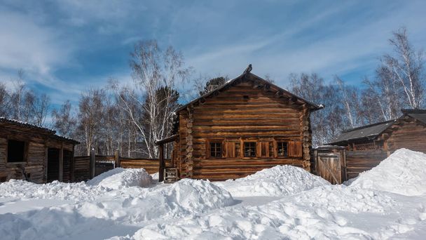 A country house made of unpainted natural logs stands among the snowdrifts. There are shutters on the small windows. Background - blue sky, white clouds, bare branches and tree trunks. - Foto, afbeelding