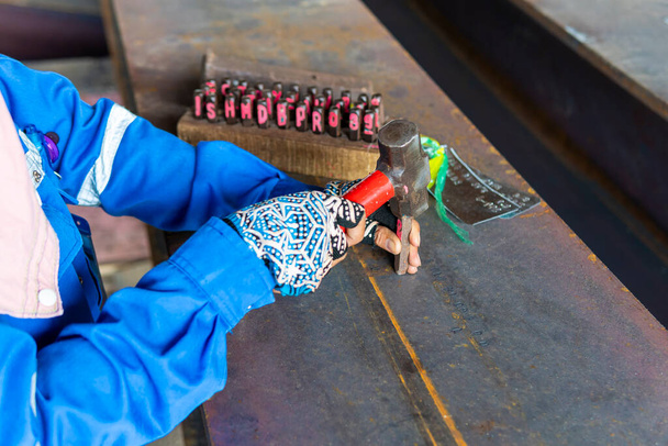 Οι εργαζόμενοι χρησιμοποιούν ένα σφυρί για να σφραγίσει τους χαρακτήρες και τον αριθμό με σφραγίδες χέρι χάλυβα στη δοκό δομή χάλυβα για την αναγνώριση κομμάτι εργασίας. - Φωτογραφία, εικόνα