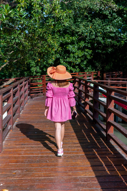 Rózsaszín ruhát viselő nő és egy narancssárga kalap sétál, hogy lássa a mangrove erdő egy fa híd sétányon. - Fotó, kép