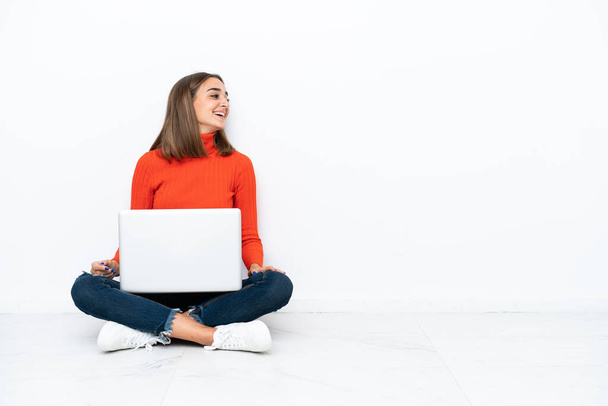 Jovem caucasiana sentada no chão com um laptop rindo em posição lateral - Foto, Imagem