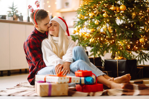 Cheerful young male and female Exchange of Christmas gifts. Young couple opening gift box near Christmas tree. Romantic day. Winter holidays. - 写真・画像