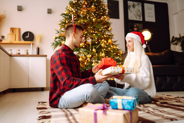 Iloinen nuori mies ja nainen Joululahjojen vaihto. Nuori pari avaamassa lahjapakkausta joulukuusen lähellä. Romanttinen päivä. Talvilomat. - Valokuva, kuva