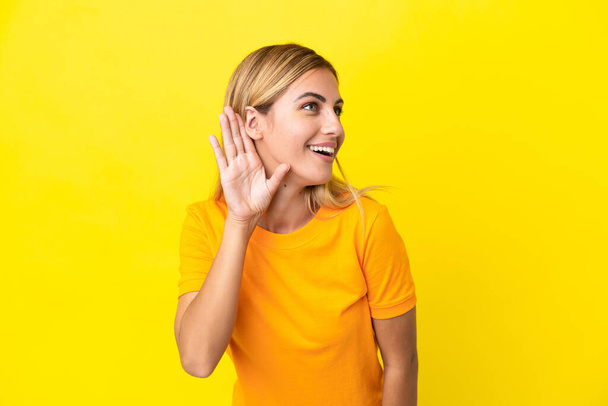 Ξανθιά Ουρουγουανή κοπέλα απομονωμένη σε κίτρινο φόντο ακούγοντας κάτι βάζοντας το χέρι στο αυτί. - Φωτογραφία, εικόνα