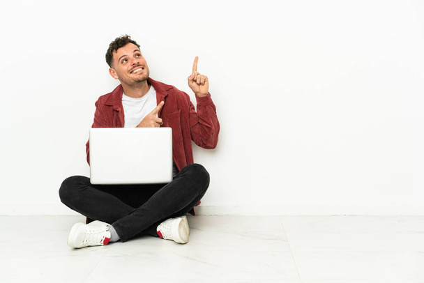 Jovem bonito caucasiano homem sit-in no chão com laptop apontando com o dedo indicador uma ótima ideia - Foto, Imagem