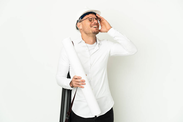 Jeune architecte homme avec casque et tenant des plans isolés sur fond blanc souriant beaucoup - Photo, image