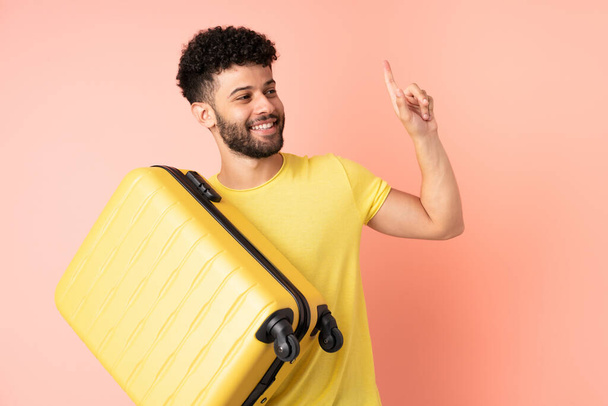 Joven hombre marroquí aislado sobre fondo rosa en vacaciones con maleta de viaje y saludo - Foto, Imagen