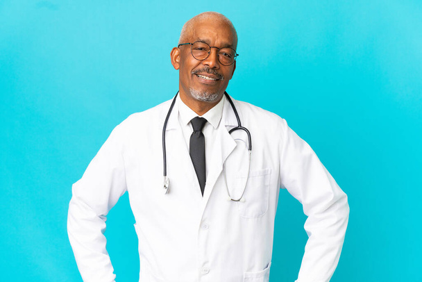 Senior arts man geïsoleerd op blauwe achtergrond poseren met armen op heup en glimlachen - Foto, afbeelding