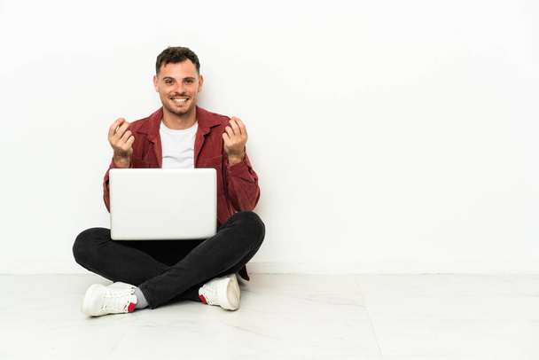 Молодой красивый белый мужчина сидит на полу с ноутбуком, зарабатывая деньги. - Фото, изображение