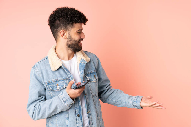 Junger marokkanischer Mann benutzt Handy isoliert auf rosa Hintergrund mit überraschendem Gesichtsausdruck, während er Seite schaut - Foto, Bild