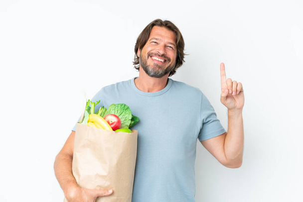 Senior holland férfi gazdaság élelmiszerbolt bevásárló táska felett elszigetelt háttér rámutatva egy nagy ötlet - Fotó, kép
