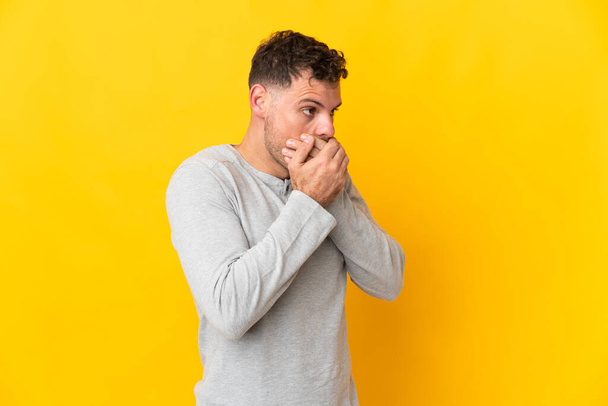 Young caucasian handsome man isolated on yellow background covering mouth and looking to the side - Foto, afbeelding