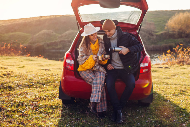 Couple amoureux se détendre dans le coffre de la voiture par le lac d'automne boire du thé à partir de la fiole à vide. Les hommes et les femmes voyagent en voiture. Les jeunes aiment les activités automnales - Photo, image