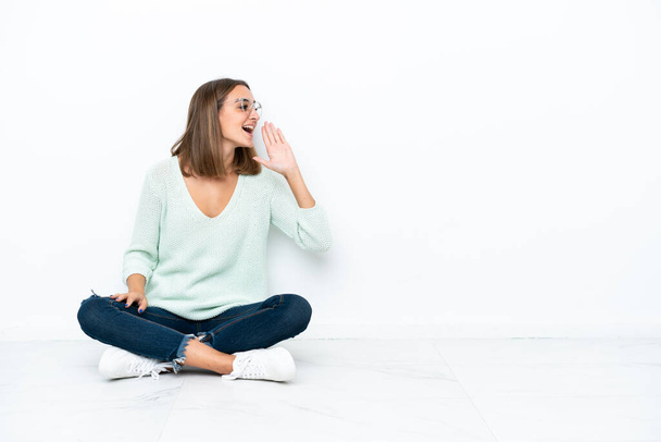 Mujer joven caucásica sentada en el suelo aislada sobre fondo blanco gritando con la boca abierta hacia el lateral - Foto, Imagen