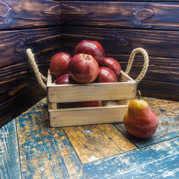 İp saplı bej bir tahta kutu olgun kırmızı elmalarla doludur. Yakınlarda, tahtaların üzerinde bir armut yatıyor. Yakın plan. - Fotoğraf, Görsel