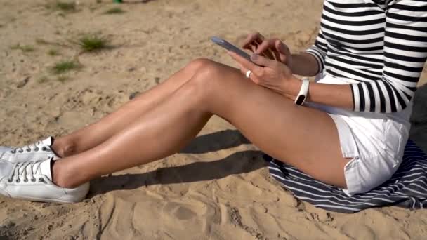 Tysiąclecia kobieta spędza czas siedząc na piasku trzymając pocztę sprawdzającą smartfona, pozytywna młoda blogerka dziewczyna influencer udostępniać treści podczas rekreacji nad morzem za pomocą telefonu komórkowego - Materiał filmowy, wideo